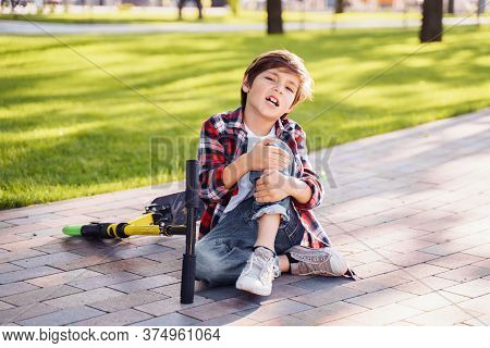 A Little Boy 8 Years Riding Scooter And Fell. Sitting On Asphalt, Holding Hands On His Injured Leg A