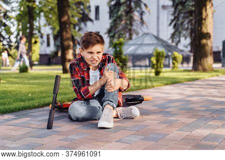 A Little Boy 8 Years Riding Scooter And Fell. Sitting On Asphalt, Holding Hands On His Injured Leg A