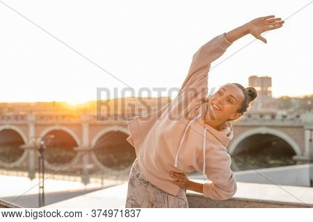 Young cheerful sportswoman in hoodie and leggins doing sidebends with her right arm stretched and raised over head during workout