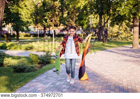 Guy Of 9 Years Old Riding Skateboard On Sunny Road In The Evening In The City Park. Holding Flutteri