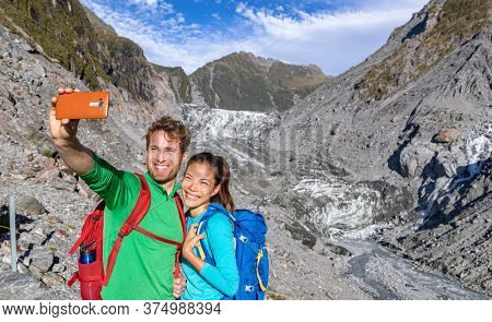 Selfie couple taking phone self-portrait on New Zealand by Franz Josef Glacier. New Zealand tourists smiling happy in nature by Franz Josef Glacier, Westland Tai Poutini National Park.