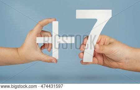 The Number Seven And The Plus Symbol In The Hands Of A Child On A Blue Background. White Number 7 Wi