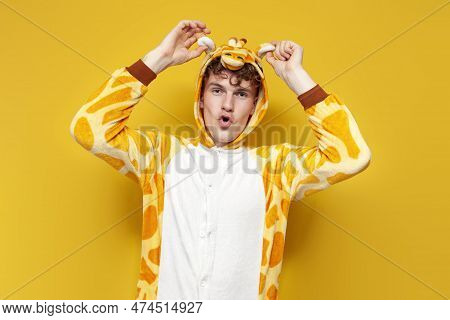 Young Joyful Guy In Funny Baby Giraffe Pajamas Grimaces And Holds His Ears On Yellow Background