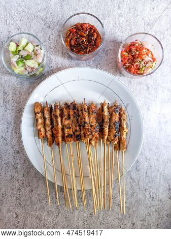 A Plate Of Chicken Satay Served With Lime, A Glass Of Pickles And Sambal Matah In The Room, Their Vi