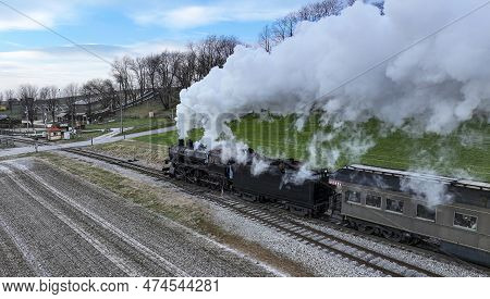 A Behind Aerial View Of A Steam Passenger Train Approaching, Traveling Thru Open Farmlands, Blowing 