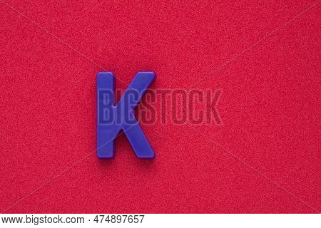 Alphabet Letter K - Violet Colored Plastic Piece On Red 032 C Foamy Background