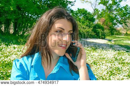 Young Woman Wearing Blue Shirt Talking With His Cell Phone Happy On The Grass With Margherites