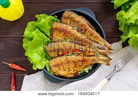 Fried fish carp (sazan) on a cast-iron frying pan with lettuce leaves on a dark wooden background. The top view.