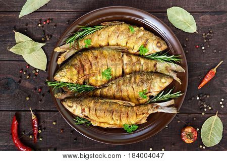 Fried fish carp (sazan) on a ceramic bowl with branches of rosemary on a dark wooden background. The top view.