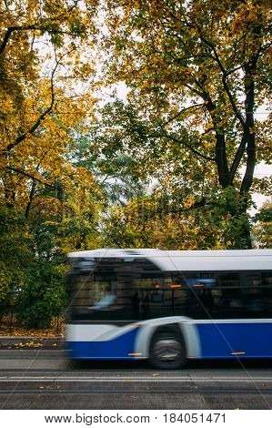 A blue public bus moves across the frame down the street with trees and foliage in the background in autumn. Abstract motion blur.