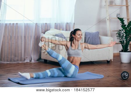 Young cheerful attractive woman practicing yoga, sitting in Sukhasana pose, working out wearing sportswear, top, pants, indoor full length