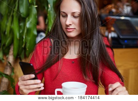 Beautiful brunette young Caucasian woman is taking rest on modern cafe bar drinking fresh morning coffee and checking her mail with modern mobile phone. Technology concept.