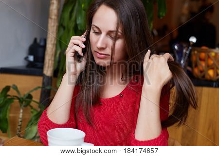 Modern organic cafe. Young beautiful lady is talking by phone with her boyfriend drinking green tea and touching her hair playfully. Health and beauty concept.