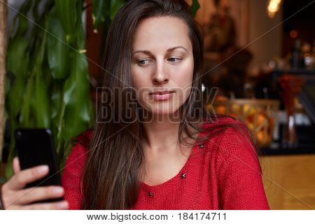 Close up shot of of beautiful young lady using her phone like a mirror or making selfie. Attractive woman is wearing red sweater sitting in a modern coffeehouse waiting for her lunch to come.