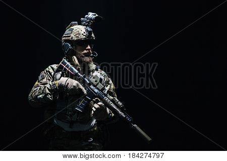 Bearded soldier in Combat Uniforms with weapon, plate carrier and combat helmet are on. Studio shot, dark background