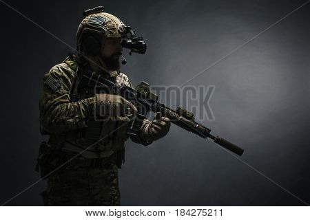 Bearded soldier in Combat Uniforms with weapon, plate carrier and combat helmet are on. Studio shot, dark background