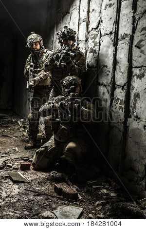 Squad of Army Rangers with rifle and machine gun moving along the concrete wall of ruined destroyed building on mission. They are ready to start firing if enemy appear. Outdoor location shot
