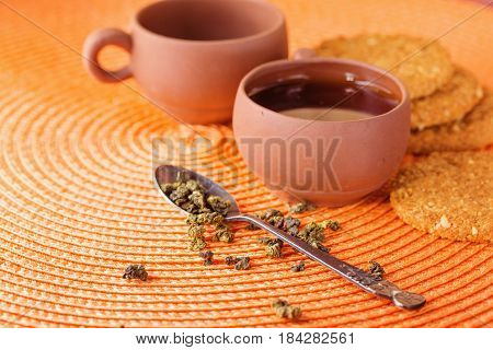 Dry leaves of green tea in a glass jar. Nearby tea leaves in a teaspoon and ceramic tea-things. Composition on a wattled napkin. The cover banks is open several tea leaves are scattered.