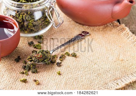 Dry leaves of green tea in a glass jar. Nearby tea leaves in a teaspoon and ceramic tea-things. Composition on a napkin from a sacking. The cover banks is open several tea leaves are scattered. Light background