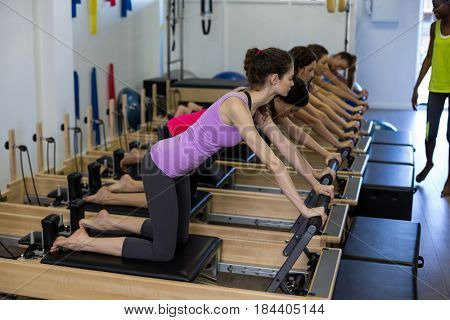 Female trainer assisting group of women with stretching exercise on reformer in gym