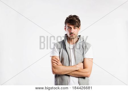 Handsome hipster fitness man in white t-hirt and gray vest with hood. Studio shot on gray background. Arms crossed.