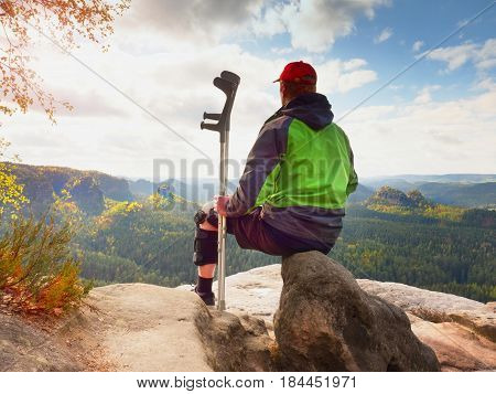 Tired Tourist With Medicine Crutch  And Broken Leg Fixed In Immobilizer Resting On Summit.