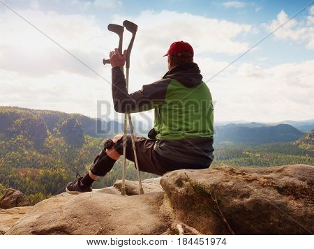 Tired Tourist With Medicine Crutch  And Broken Leg Fixed In Immobilizer Resting On Summit.