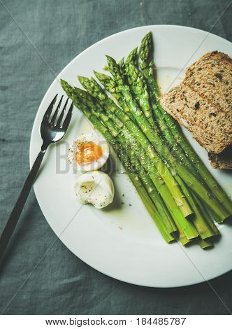 Cooked asparagus with soft-boiled egg, grilled bread and herbs on white round plate over grey textile background, top view. Clean eating food concept