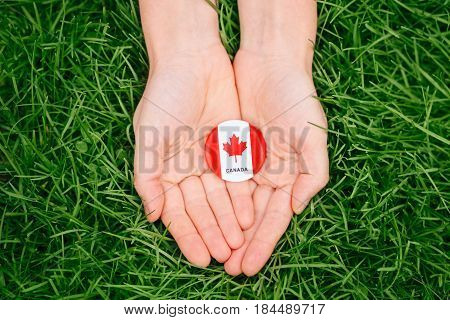 Macro closeup shot of hands palms holding round badge with red white canadian flag maple leaf on green grass forest nature background outside Canada Day celebration