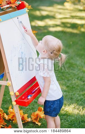 White Caucasian toddler child kid girl standing outside in summer autumn park drawing on easel with markers looking away playing studying learning back to school concept