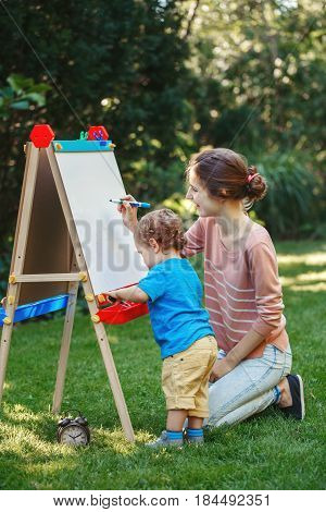 White Caucasian toddler child kid girl with older sister teacher standing outside in summer autumn park drawing on easel with markers playing studying learning back to school