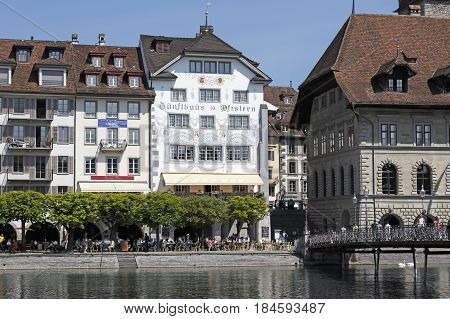 Lucerne Switzerland - May 08 2016: Houses by the riverside and the alley along the shore which is planted with trees under which is an outdoor seating restaurant there are also some people.