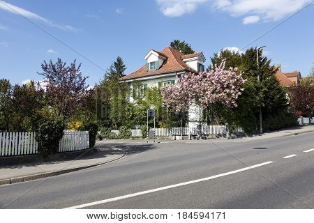 Bern Switzerland - April 14 2017: Residential houses hidden among the lush vegetation of many bushes and trees on a narrow street in a quiet district. White fences of the property can be seen.