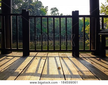 Wooden railing shadow casting on vintage balcony floor of water front pavilion with a little far off forest and lake. Afternoon sun light and hard shadows.