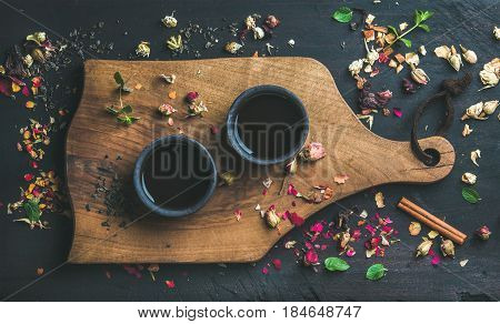 Chinese black tea in black stoneware cups on serving wooden board over black wooden background with herbs, flower buds, tea leaves spilt around, top view