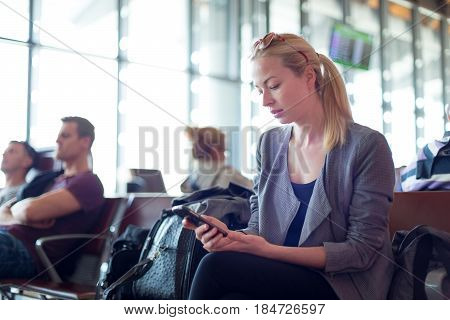 Casual blond young woman using her cell phone while waiting to board a plane at the departure gates. Wireless network hotspot enabling people to access internet conection. Public transport.