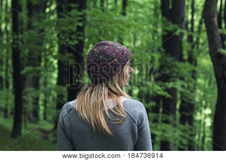 horizontal rear view portrait of Caucasian young woman with long blonde hair and colorful wool hat standing in a green forest