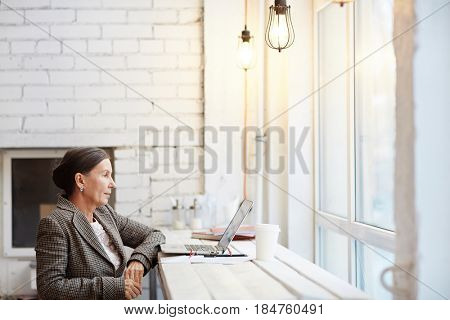 Profile view of beautiful mature office worker preparing presentation on laptop while sitting in cyber cafe with paper cup of cappuccino, waist-up portrait