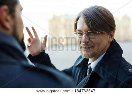 Bearded middle-aged manager looking at his colleague with squinty eyes while talking to him outdoors, over shoulder view
