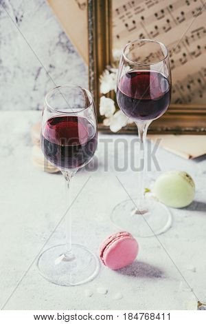 Two glasses of port wine with variety of colorful french dessert macaron macaroons with different fillings served with spring flowers and old music notes over gray texture background.