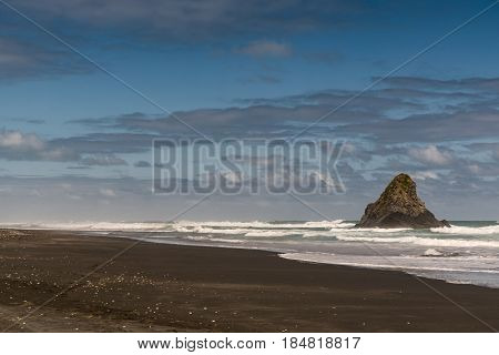 Auckland New Zealand - March 2 2017: Black volcanic sand Karakare Beach under blue cloudy sky with Te Kaka Whakaara Rock (Watchman) in the surf of Tasman Sea.