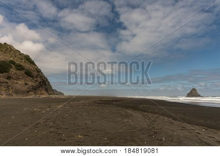 Auckland New Zealand - March 2 2017: Wide shot of black sand Karakare Beach under blue cloudy sky with Te Kaka Whakaara Rock (Watchman) in the surf of Tasman Sea far away. Rocky cliff.