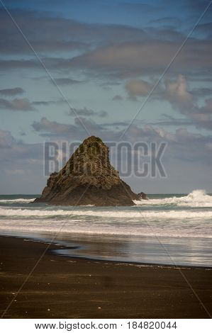 Auckland New Zealand - March 2 2017: Portrait of black sand Karakare Beach under blue cloudy sky with Te Kaka Whakaara Rock (Watchman) in the surf of Tasman Sea.