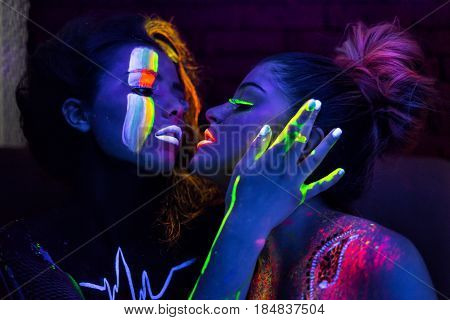 Sexy lesbian fashion models in uv neon light with fluorescent glowing Body Art make-up kissing. Low key dark image. Soft focus image.