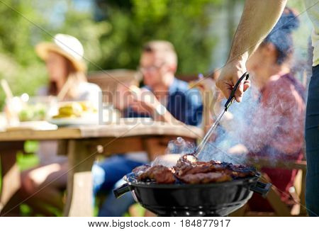 leisure, food, people and holidays concept - man cooking meat on barbecue grill for his friends at summer outdoor party