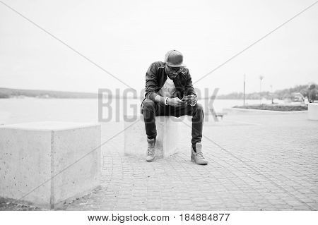 Portrait Of Stylish African American Man On Sportswear, Cap And Glasses Sitting At Stone Cube With P