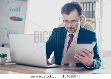 Serious Worker Is Typing Information From Tablet To Laptop. He Is In Formalclothes, Wearing Glasses,