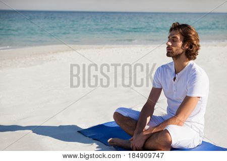 Man with eyes closed meditating while sitting on exercise mat at beach