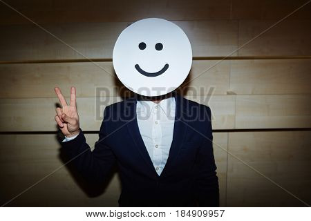 Waist-up portrait of successful entrepreneur in formalwear standing against wooden wall and showing V sign at camera, his face covered with smiling mask