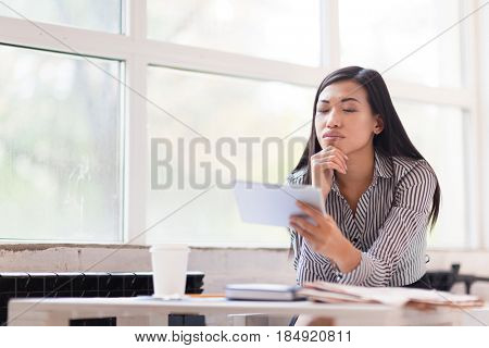 Pensive Asian entrepreneur rubbing her chin while analyzing statistic data displayed on screen of digital tablet, waist-up portrait